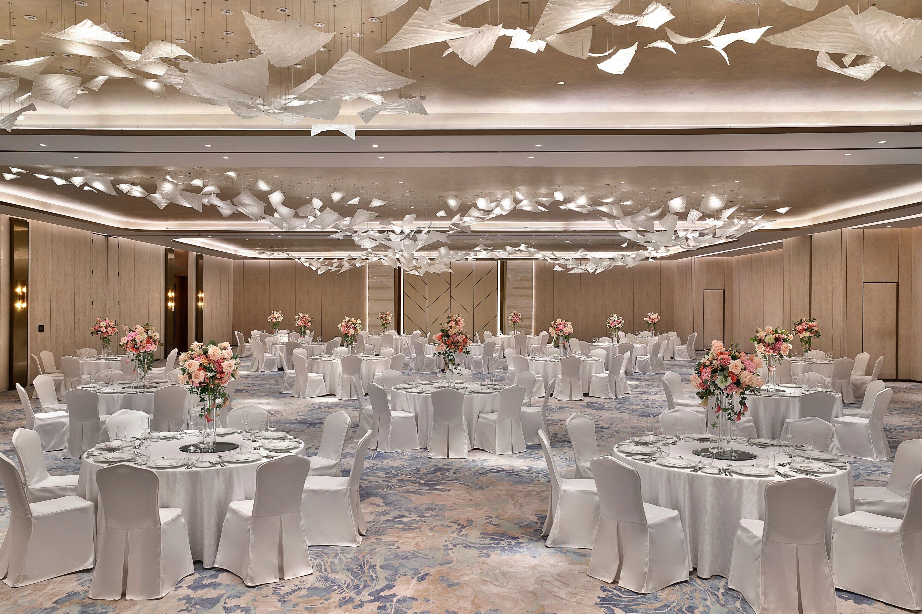 Un amplio salón de banquetes elegantemente decorado con mesas redondas, sillas blancas y centros de mesa florales.