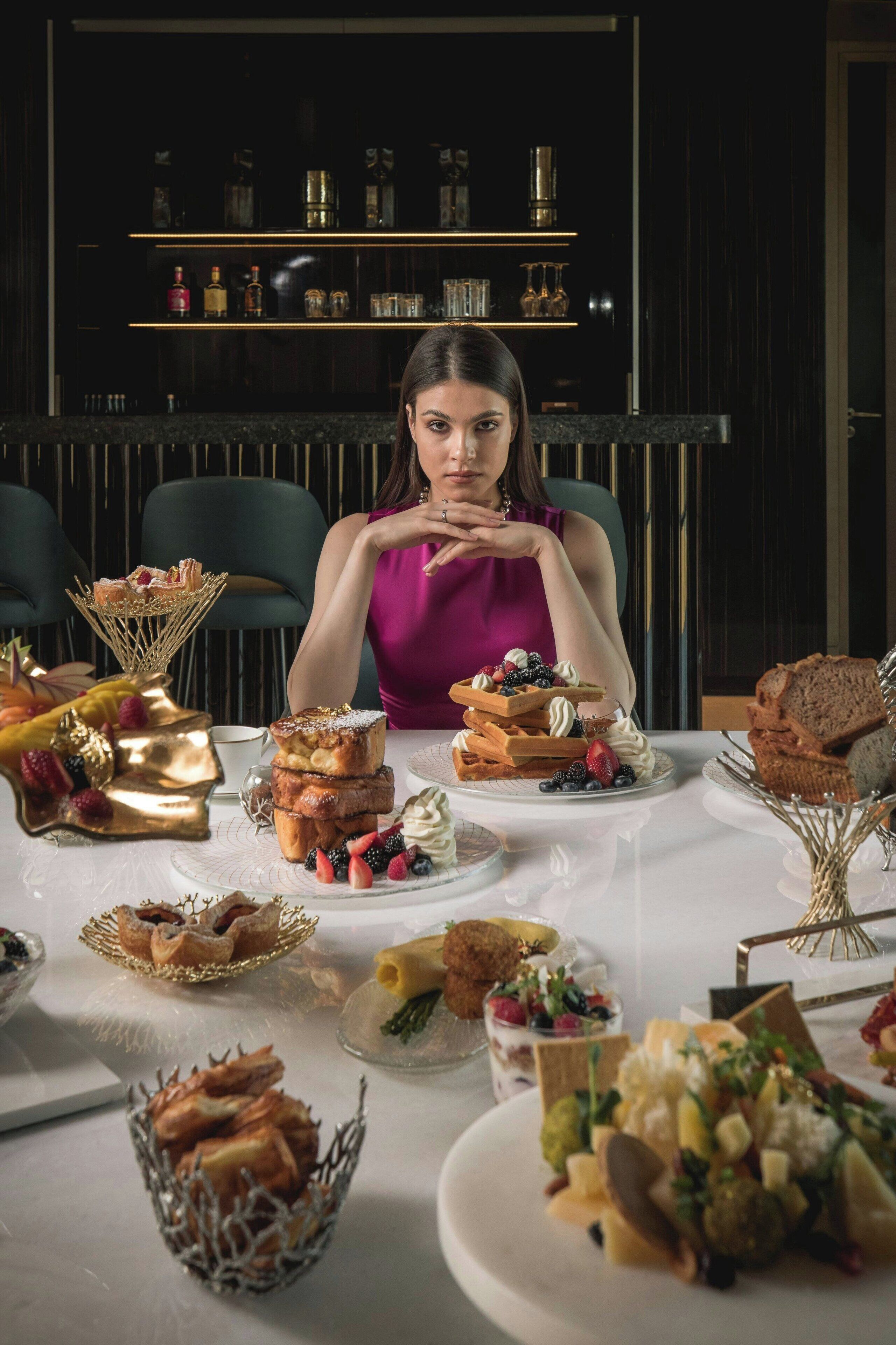 Mesa posta com diversos doces e sobremesas, uma pessoa sentada à mesa e um bar ao fundo, com estantes.