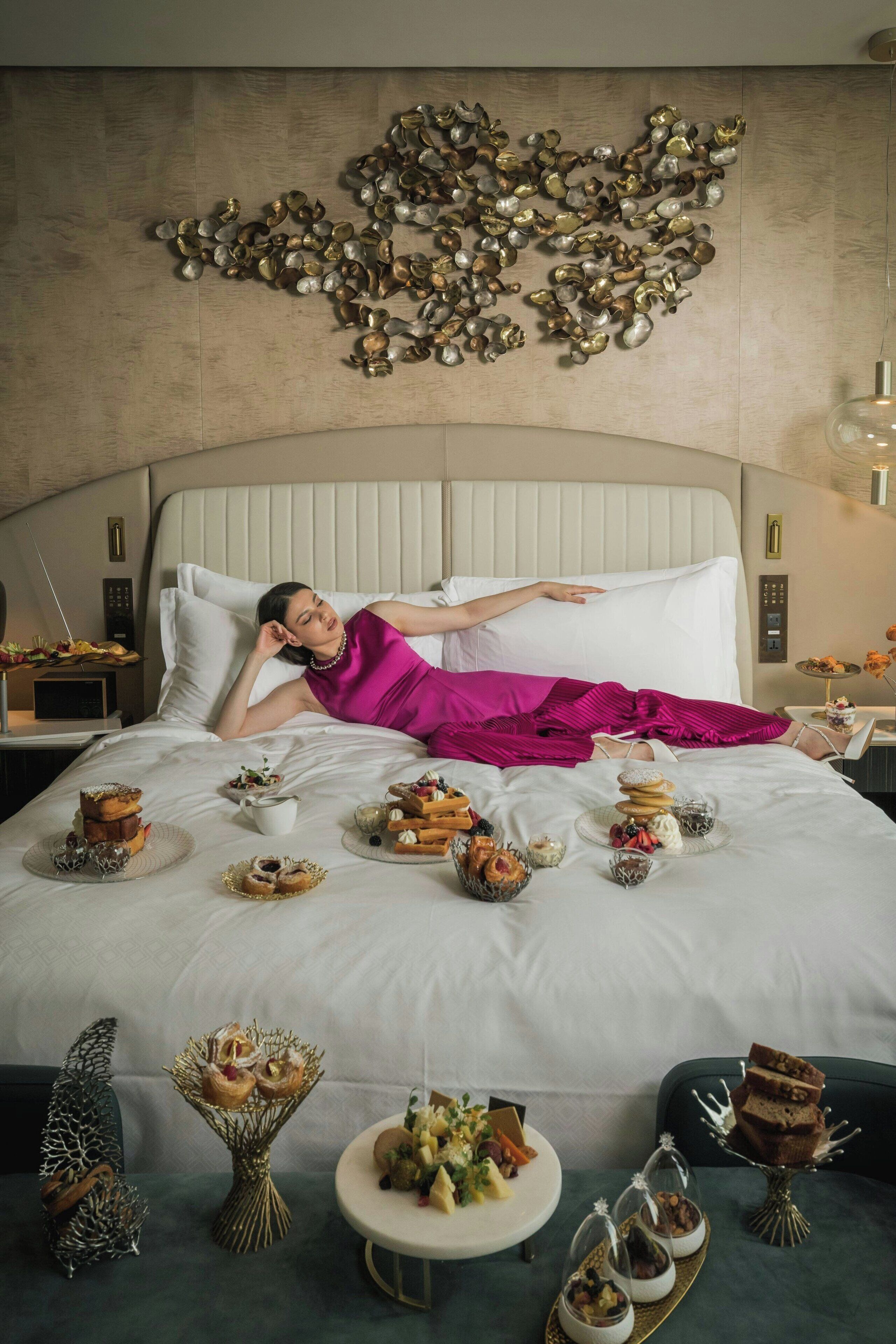 A woman lying on a bed with a breakfast spread, including pastries and desserts, in a room with a decorative wall and a nightstand with a lamp.