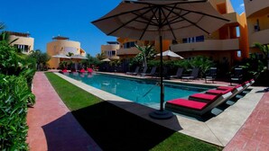 Piscina con sillas de descanso y sombrillas, rodeada de vegetación y edificios de varios pisos.