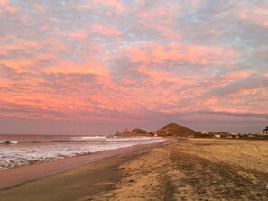 A beach with a sunset, waves, and buildings in the distance. - Modern Cerritos Home • Walk to Beach & Dining (El Pescadero)