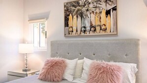 A bedroom with a grey upholstered headboard, a bed with white bedding and two fluffy pink pillows, a bedside table with a lamp, and a wall-mounted artwork of surfboards.