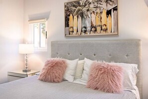 Et soveværelse med en grå polstret sengegavl, en seng med hvide sengetæppe og to bløde pink puder, et natbord med en lampe, og et billede af surfboards på væggen.