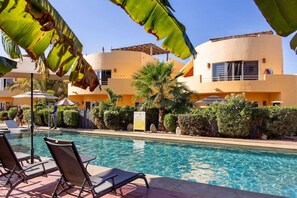 A poolside area with lounge chairs and a clear pool, surrounded by palm trees and a building with a yellow facade. - Modern Cerritos Home • Walk to Beach & Dining (El Pescadero)