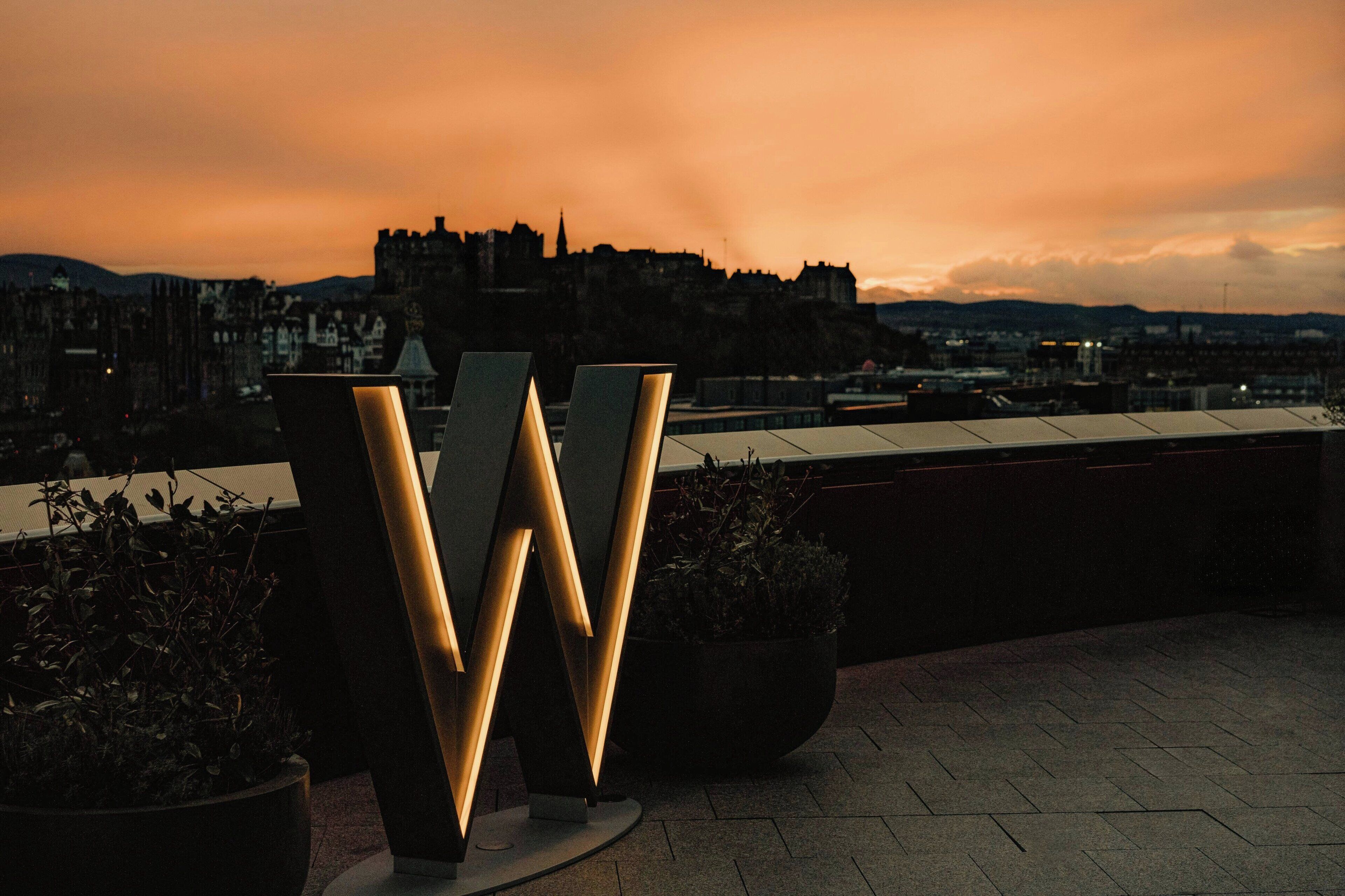 A rooftop terrace with modern, illuminated letter-shaped sculptures, potted plants, and a view of a historic castle in the background.