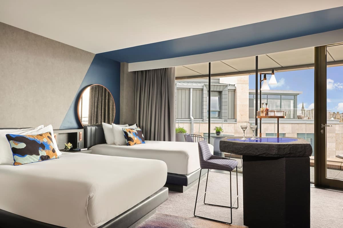 A hotel room with two beds, a small bar area, and a view of the cityscape.