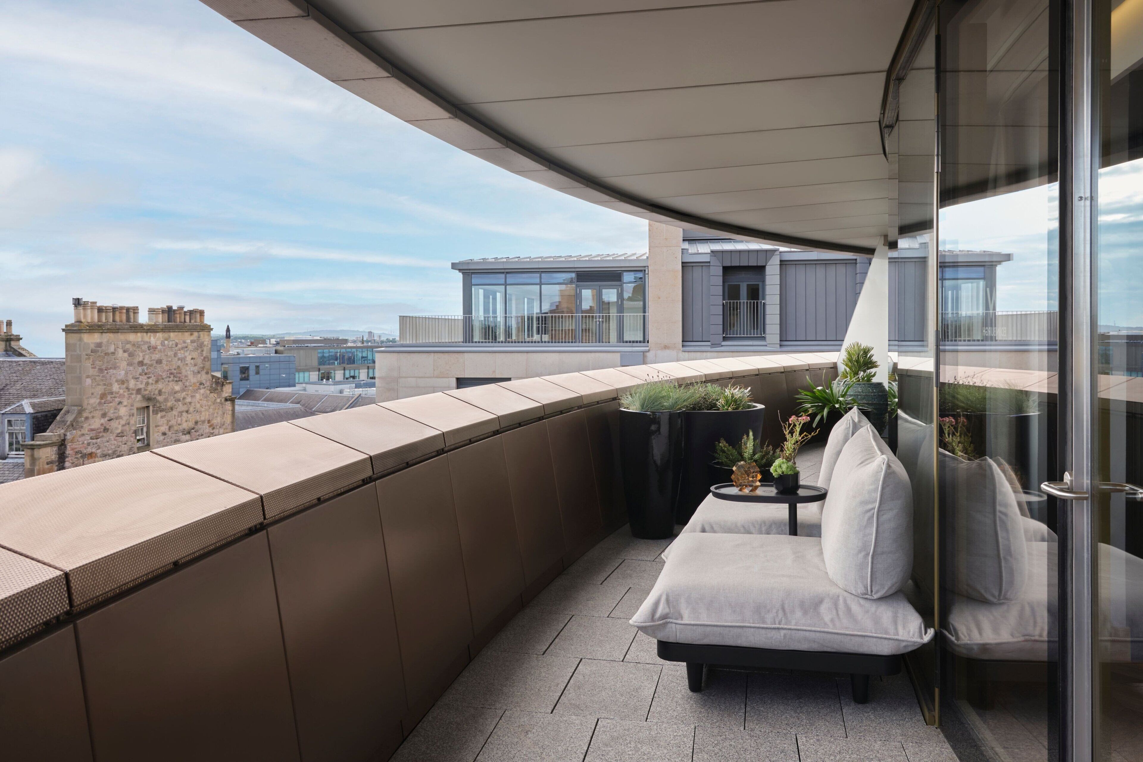 A modern balcony with a sofa, potted plants, and a glass railing overlooking a cityscape.