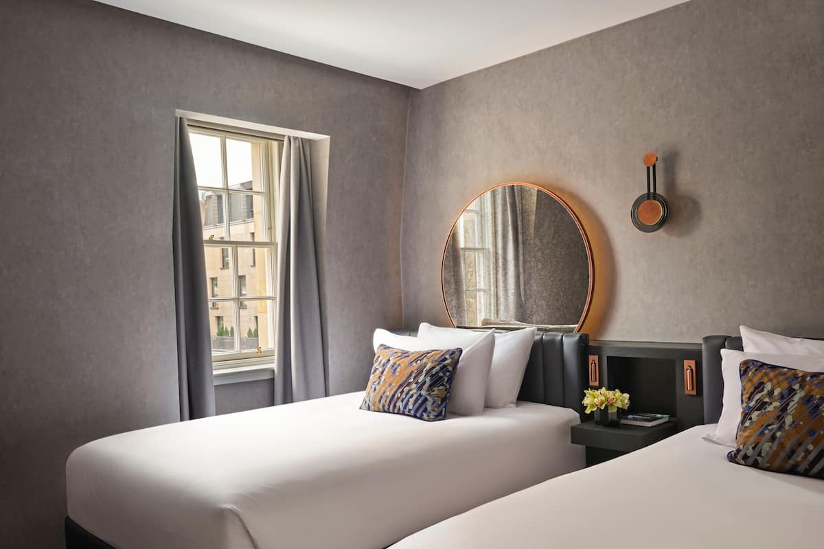 A hotel room with two beds, a large mirror, a small table with a vase, and a window with curtains.