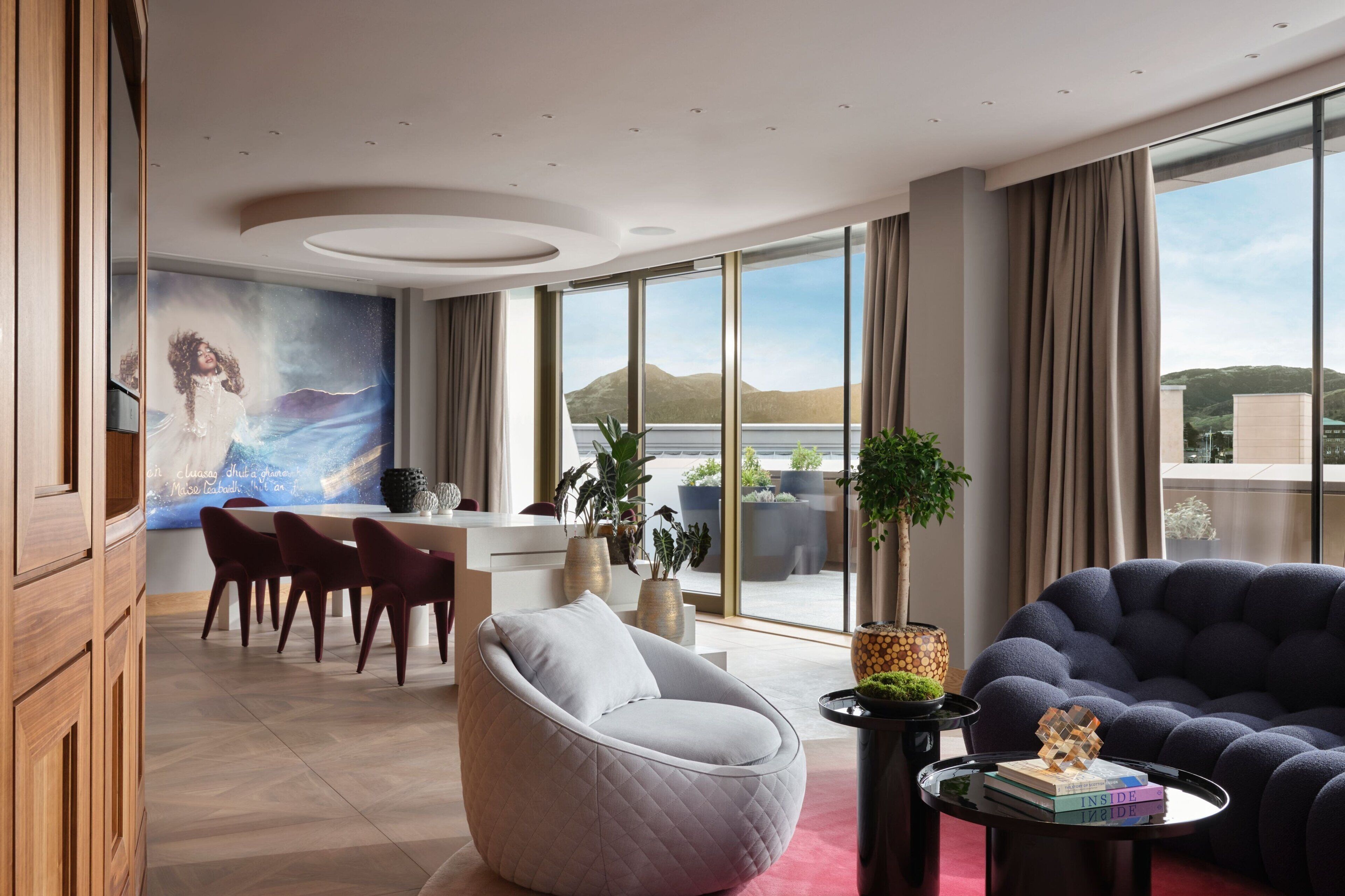 Una habitación de hotel moderna con comedor, un ventanal con vistas a las montañas y una zona de estar acogedora con una mesa redonda y sillas.
