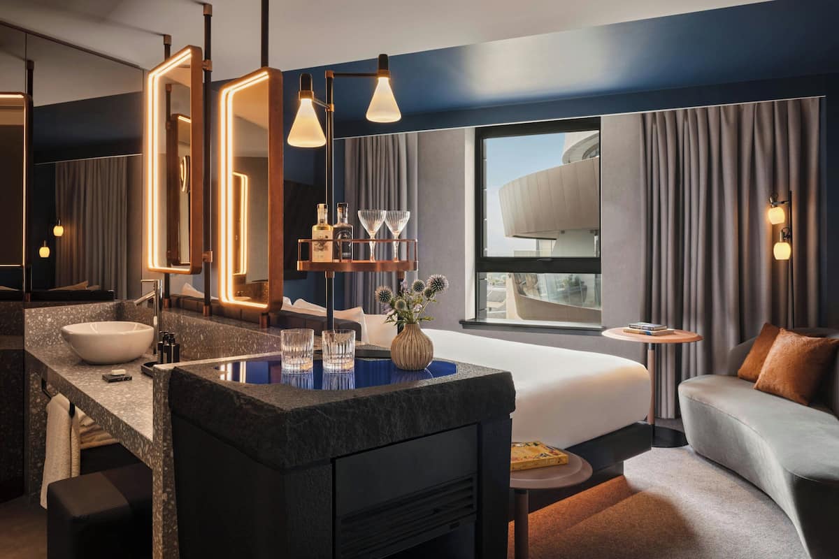 A modern hotel room with a large bed, a sitting area, a vanity with a large mirror, and a view of the city.