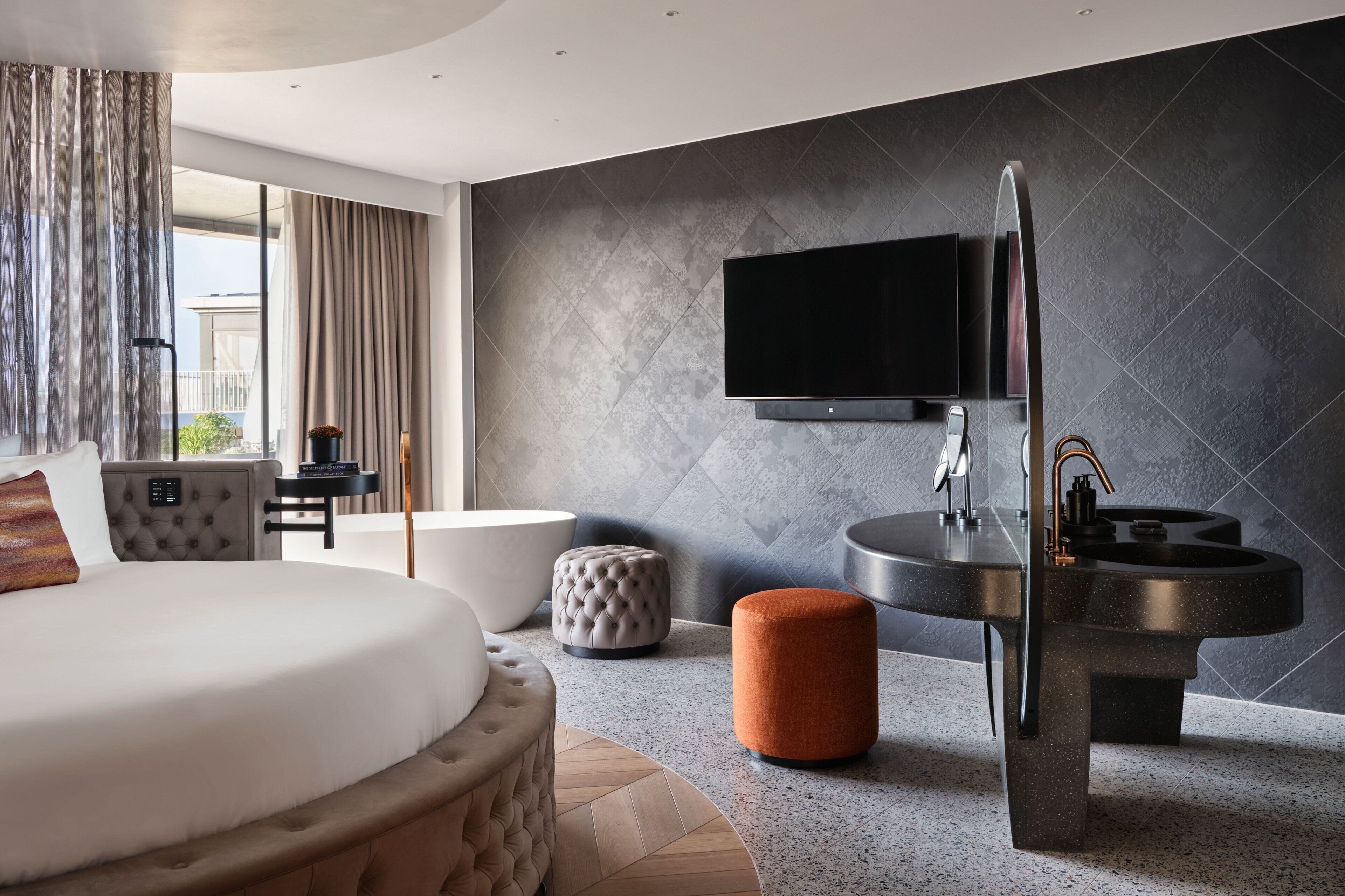 Una habitación de hotel moderna con una cama grande, un televisor de pantalla plana montado en la pared, una mesa de centro redonda y un diseño de lavabo original.
