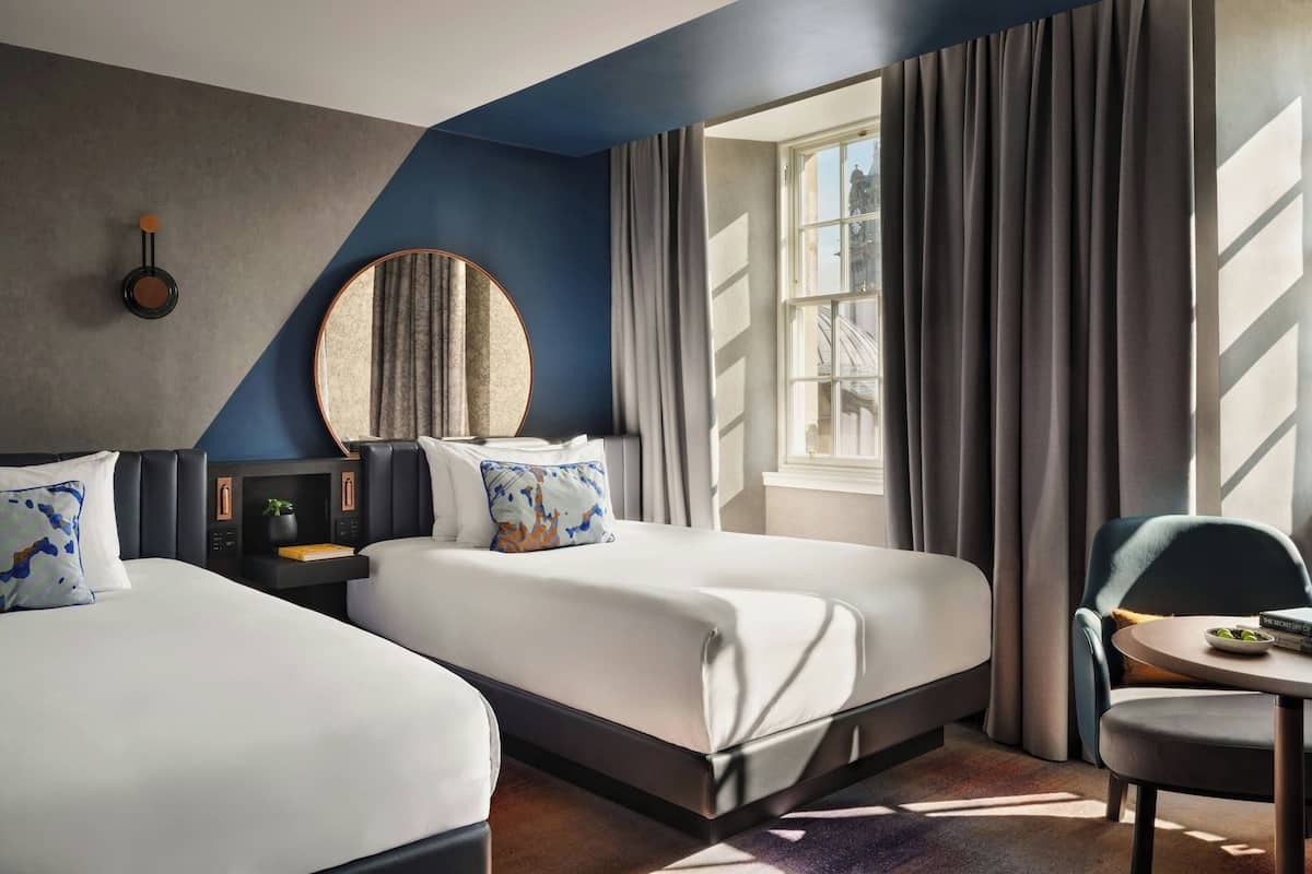 A hotel room with two beds, a round mirror, a small table, and a chair.