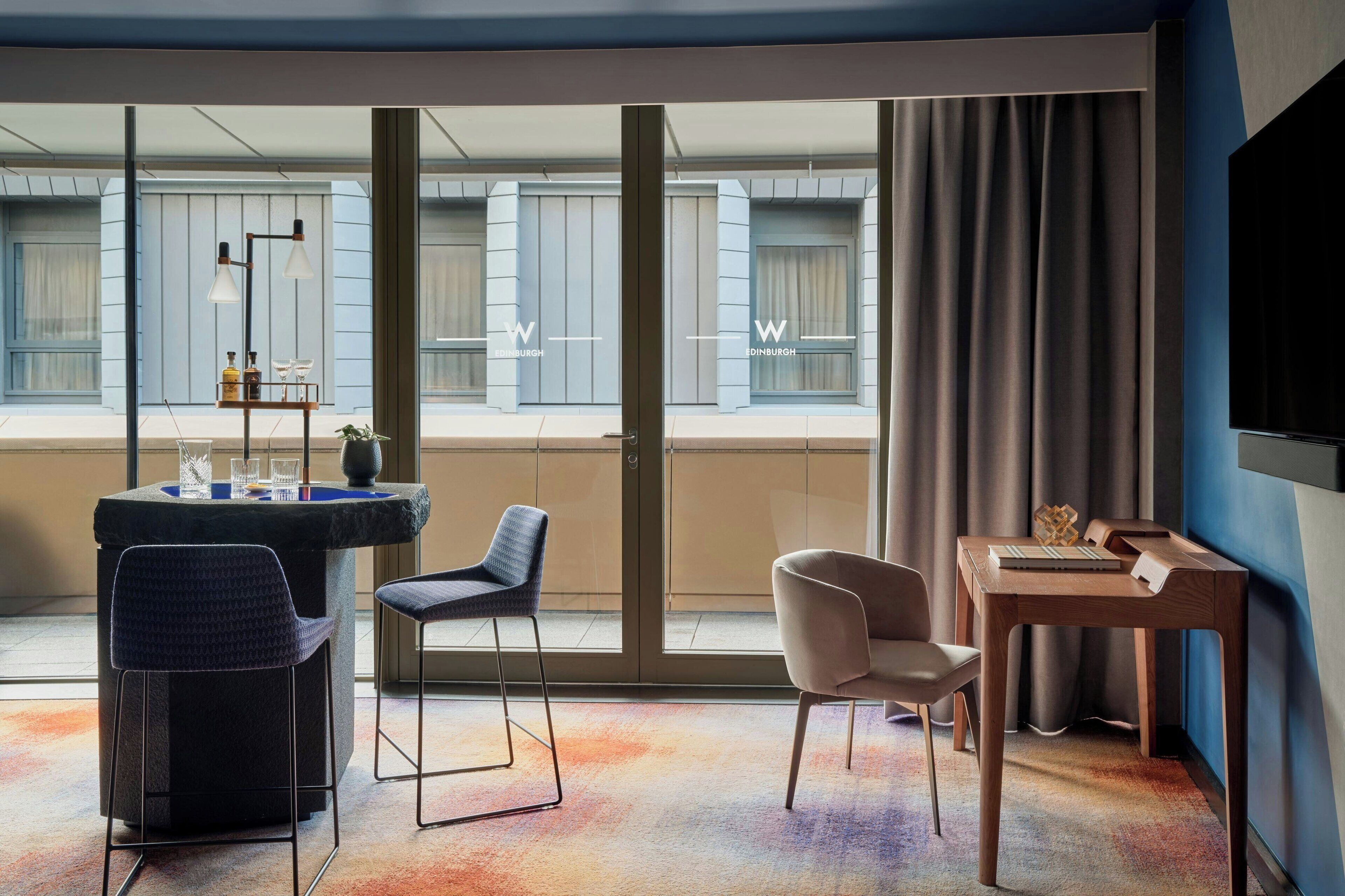 Una habitación de hotel moderna con un ventanal grande, un escritorio con computadora, una silla y una mesita con una botella y vasos.