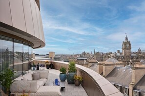 A rooftop terrace with a curved glass wall, a sofa, potted plants, and a view of historic buildings in the city.