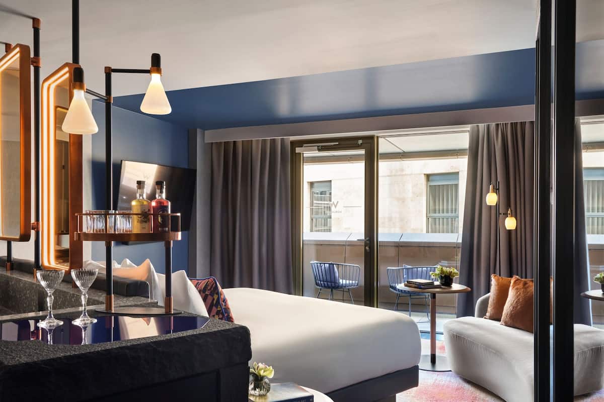 A modern hotel room with a large bed, a sitting area with a sofa and armchairs, a small table with a vase, and a balcony with a view of the city.