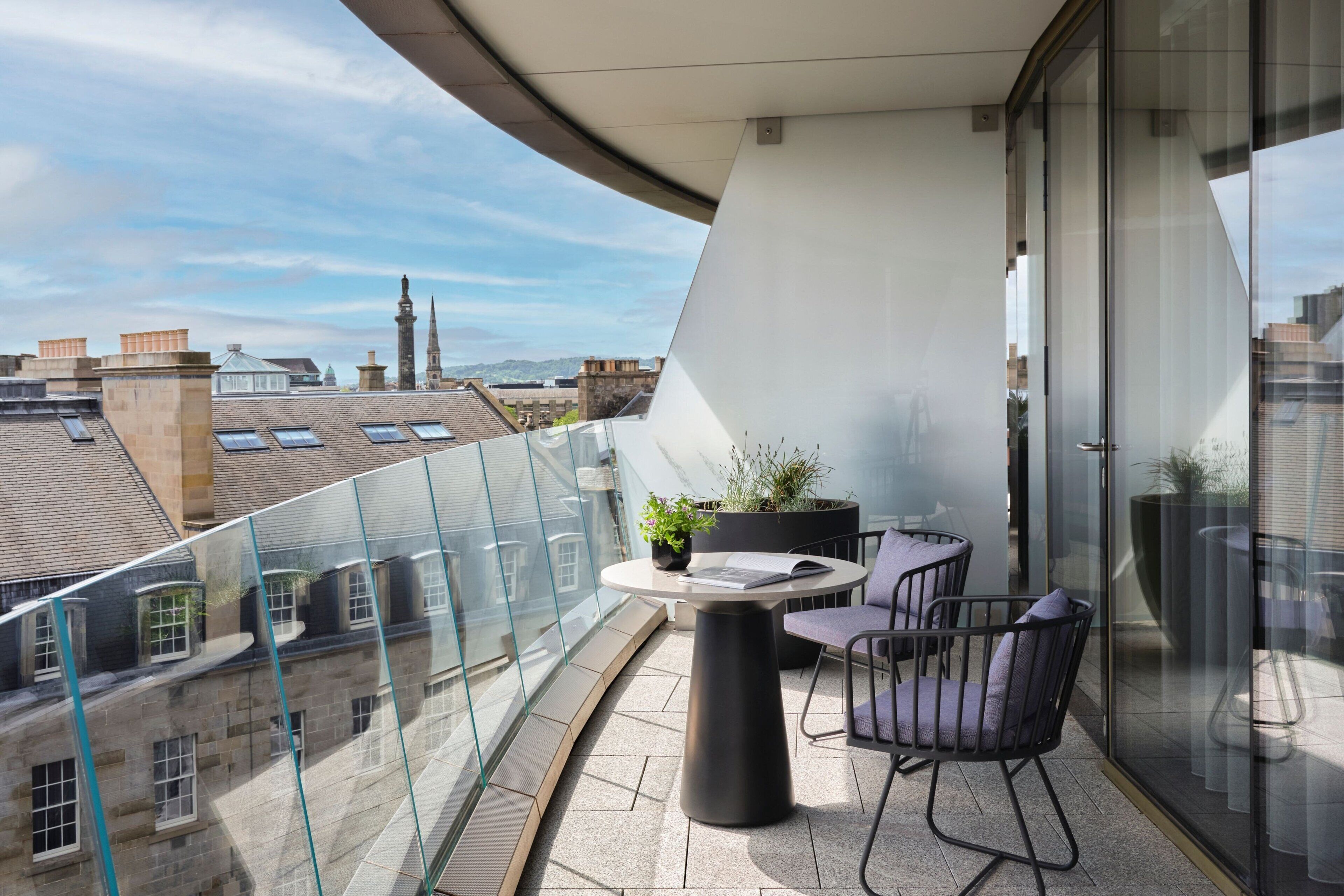 A balcony with a glass railing, a round table, two chairs, and potted plants.