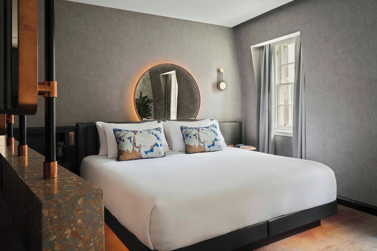 A modern hotel room with a large bed, a round mirror, a nightstand, and a window with curtains.