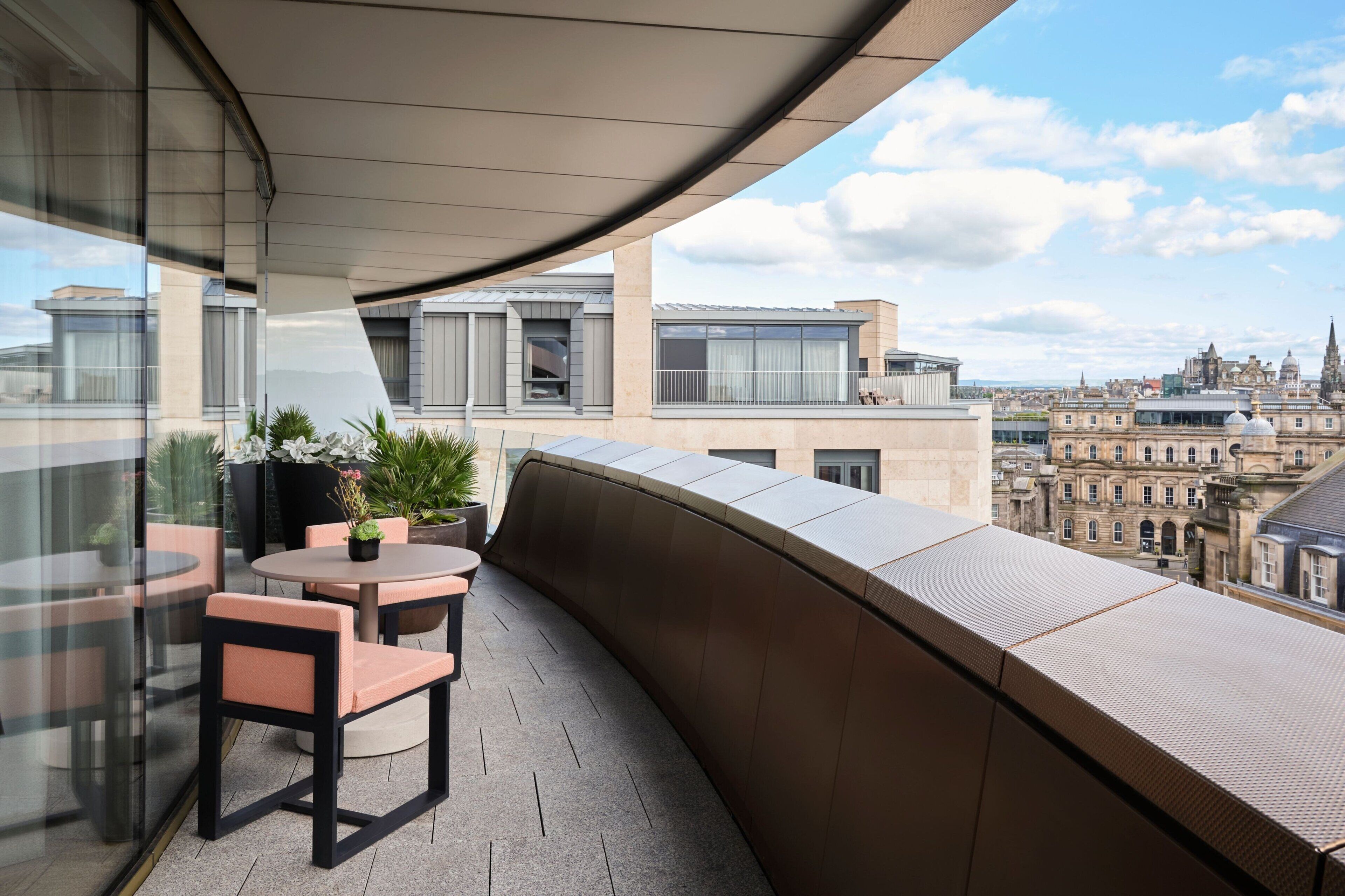 A modern balcony with a curved glass wall, a round table, and chairs, overlooking a cityscape with historic buildings.