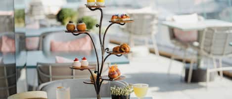 Una mesa de comedor con un expositor escalonado de postres, un vaso de agua, una bebida y una vaporera de bambú con dim sum.