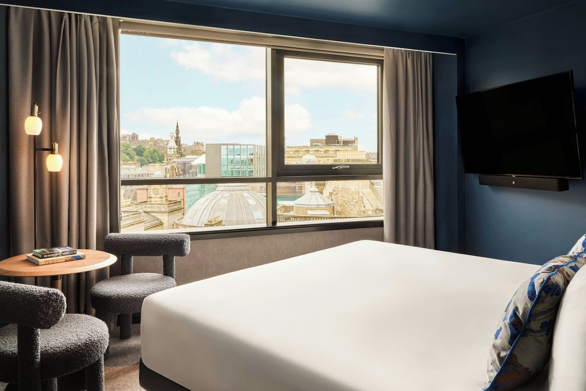 Una habitación de hotel con una cama grande, un televisor montado en la pared, una pequeña mesa redonda con dos sillas y vistas a la ciudad a través de un amplio ventanal.