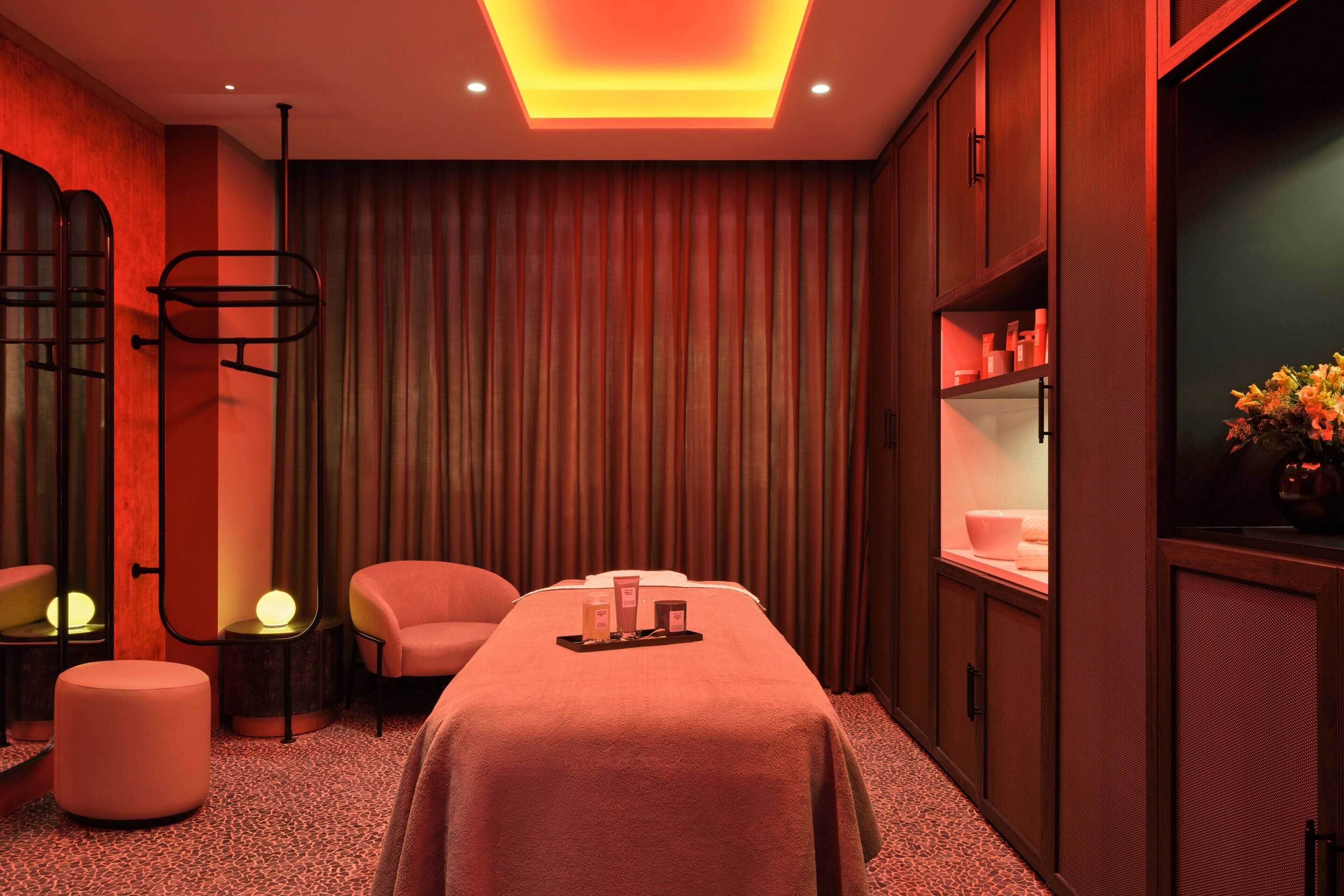 A spa room with a massage table, two chairs, a small table with toiletries, and a shelf with bottles.