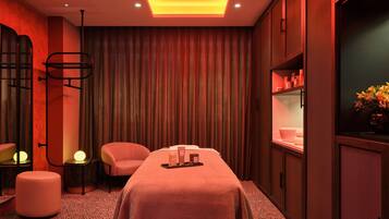 A spa room with a massage table, two chairs, a small table with toiletries, and a shelf with bottles.