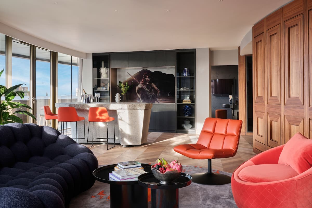 A modern living room with a sofa, a coffee table with books and fruit, and a red chair. There is a kitchen area with a large island and a dining area with red chairs.