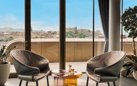 A balcony with two leather chairs, a glass coffee table, and a view of a cityscape.