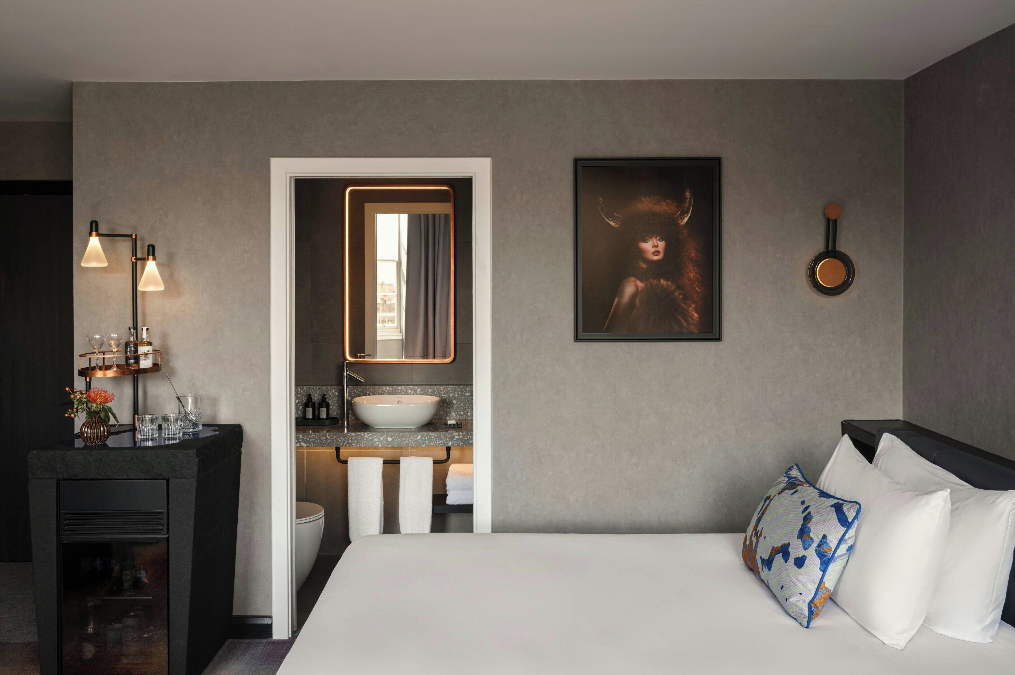 A hotel room with a bed, a bedside table with a lamp, a bathroom with a sink and mirror, and a framed picture on the wall.