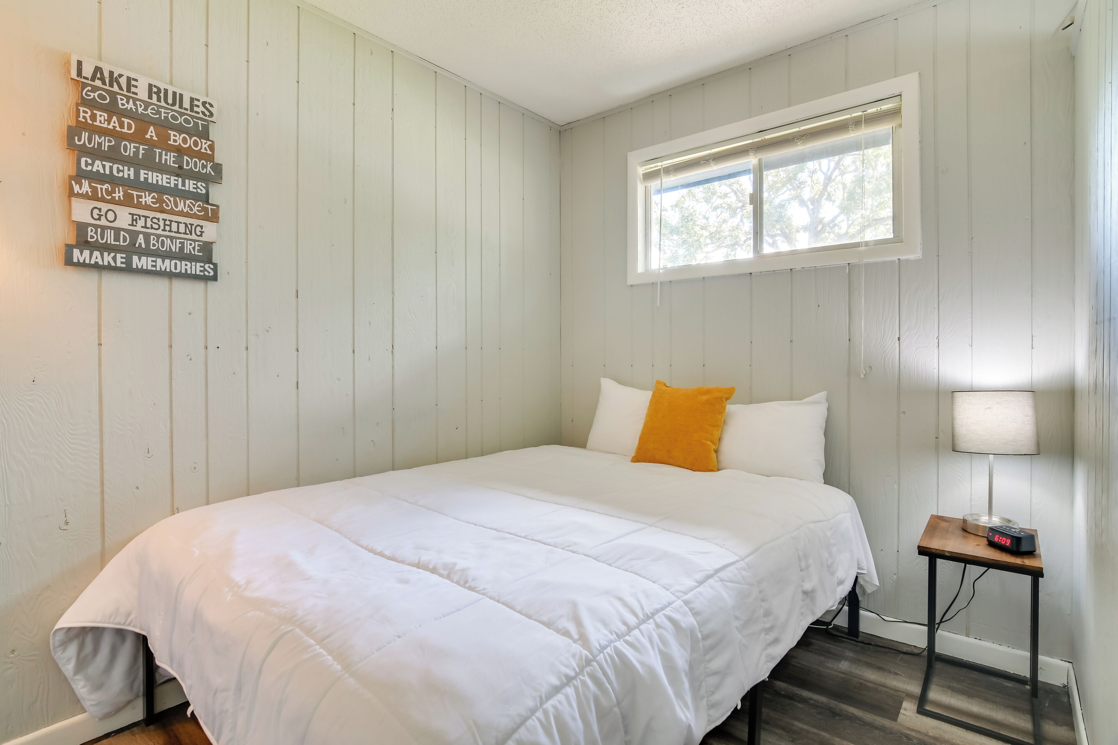 Un dormitorio con cama, ventana, mesita de noche con lámpara y un cartel en la pared que enumera actividades.