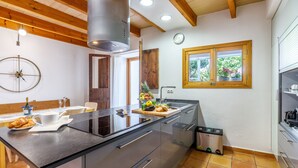 A modern kitchen with a large island, wooden ceiling beams, a window with a view of greenery, and a clock on the wall.