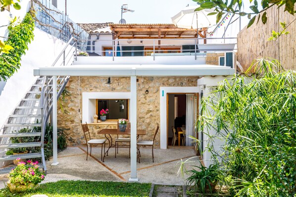 Una casa de dos pisos con fachada de piedra, una pérgola de madera, un área de comedor con mesa y sillas, y una escalera que conduce al piso superior.