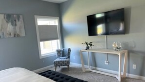 Un dormitorio con cama, una silla, un mueble consola con televisor y una ventana con persianas.