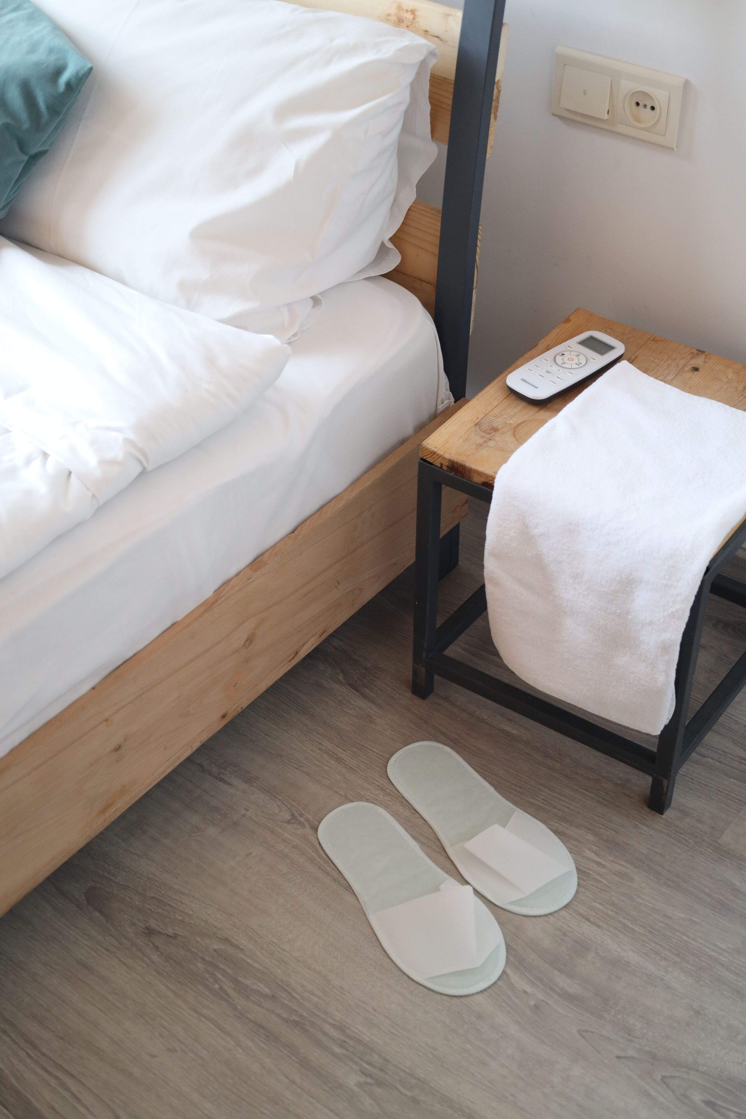 A neatly made bed with white linens, a wooden nightstand with a white towel and a digital device, and a pair of slippers on the floor.