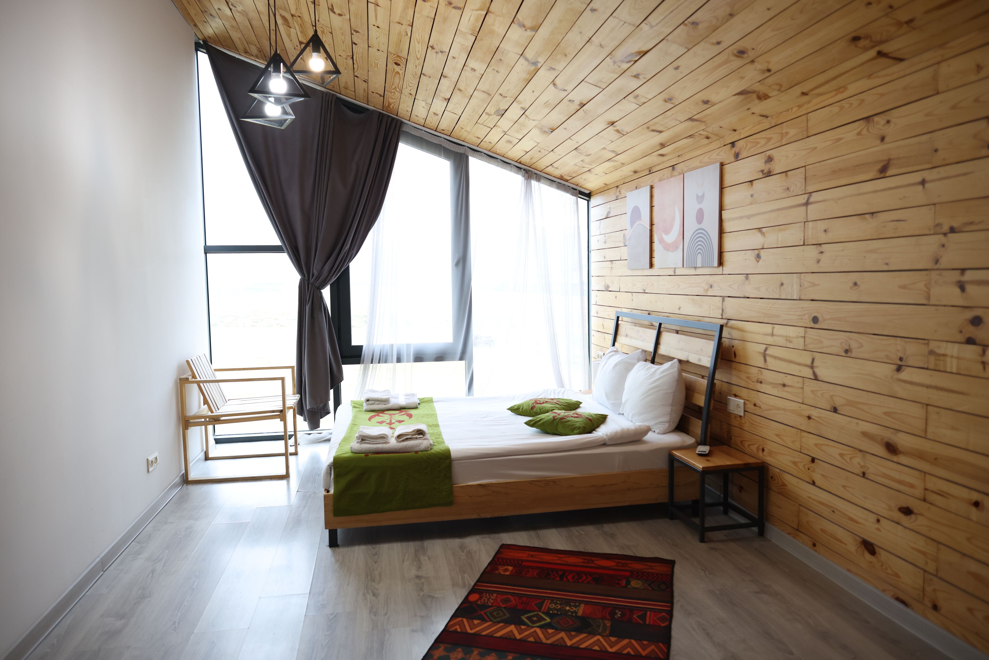 A bedroom with a wooden ceiling, a bed with pillows, a bench, and a window with curtains.