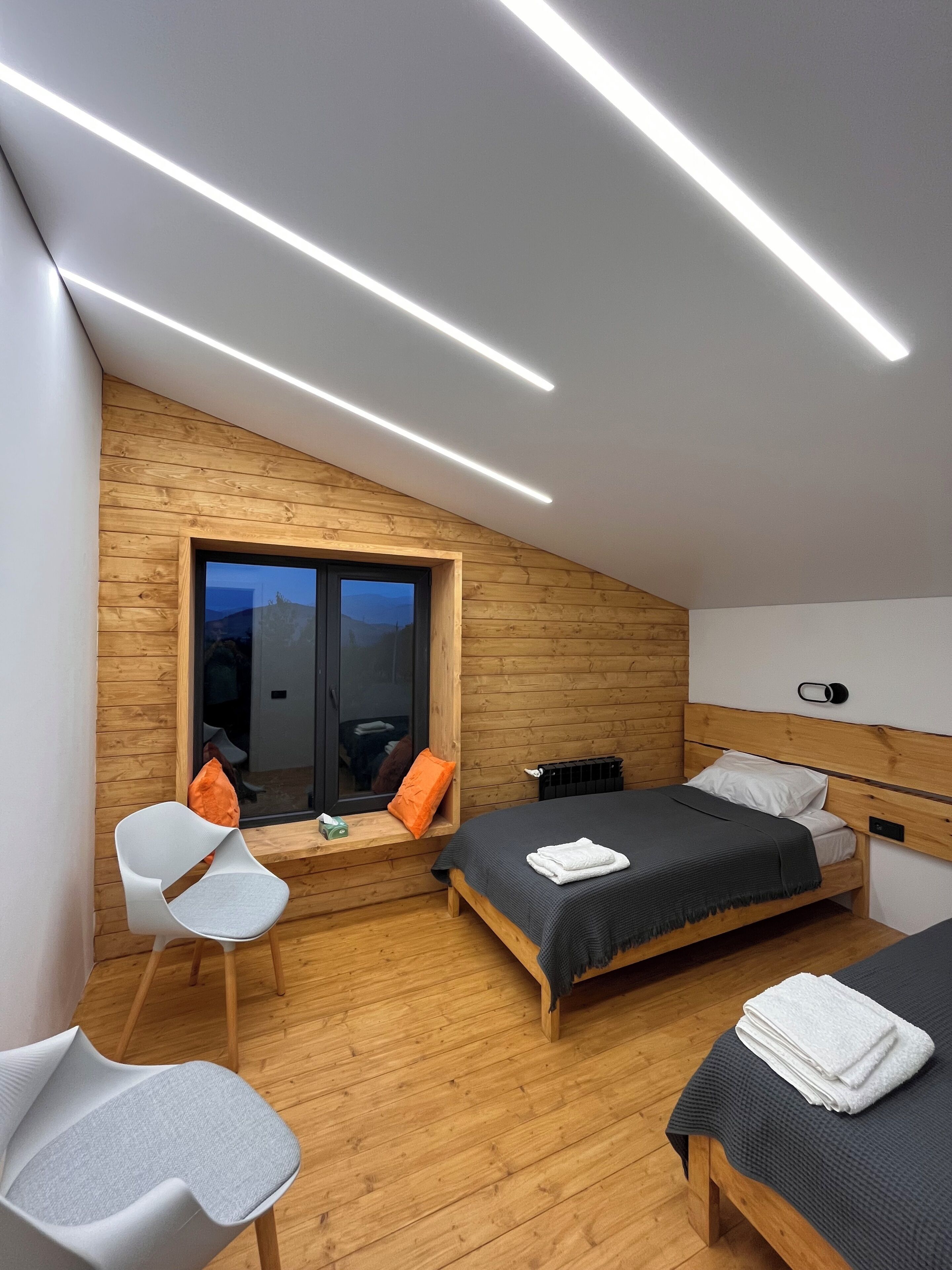 A modern bedroom with a sloped ceiling, wooden walls, a large window, a bed with a dark bedspread, a white chair, and a bedside table with a cup.