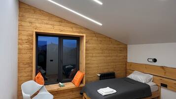 A modern bedroom with a sloped ceiling, wooden walls, a large window, a bed with a dark bedspread, a white chair, and a bedside table with a cup.