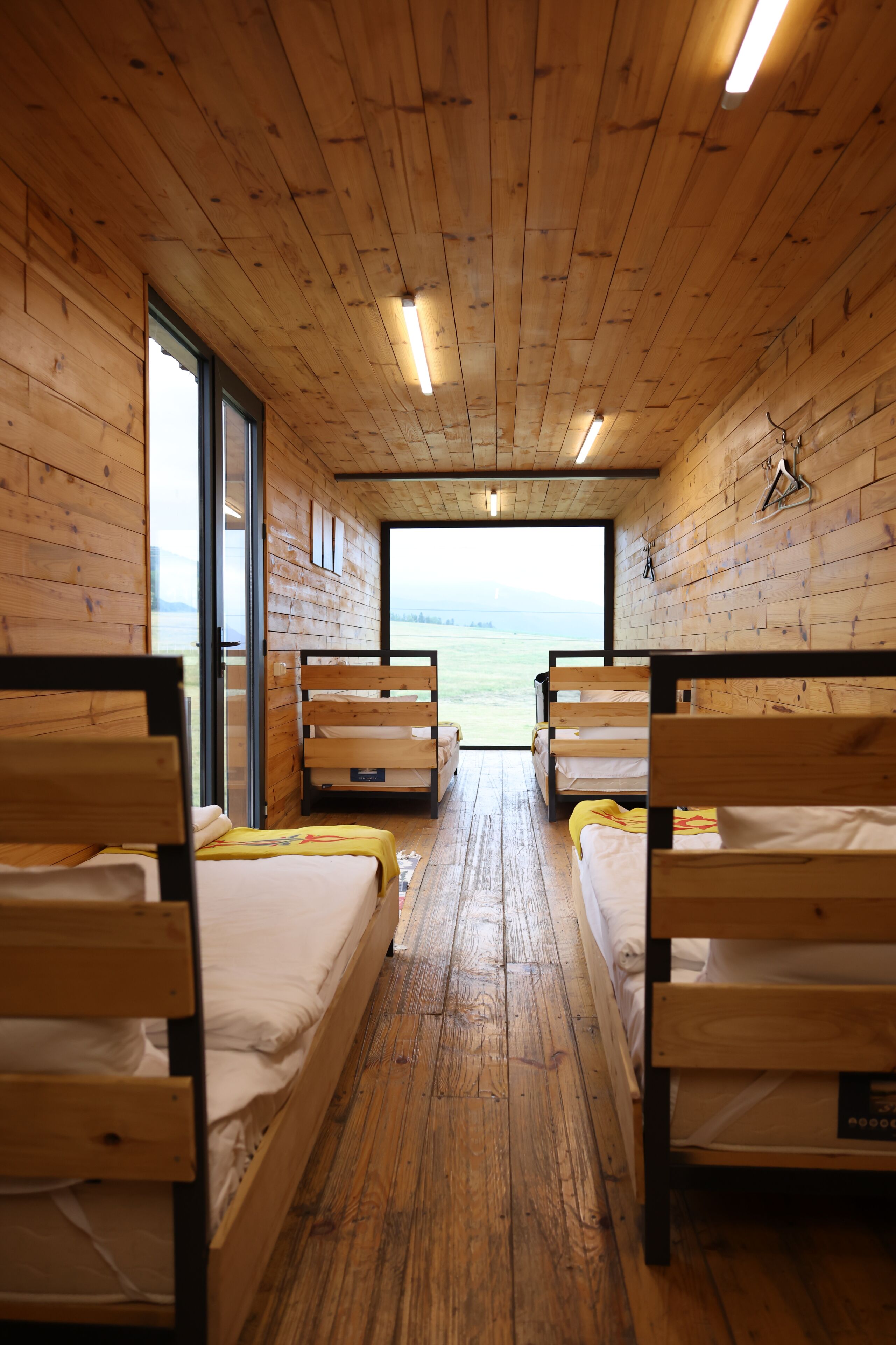 A cabin with wooden walls and floors, two beds, a bench, and a window with a view of the sea.