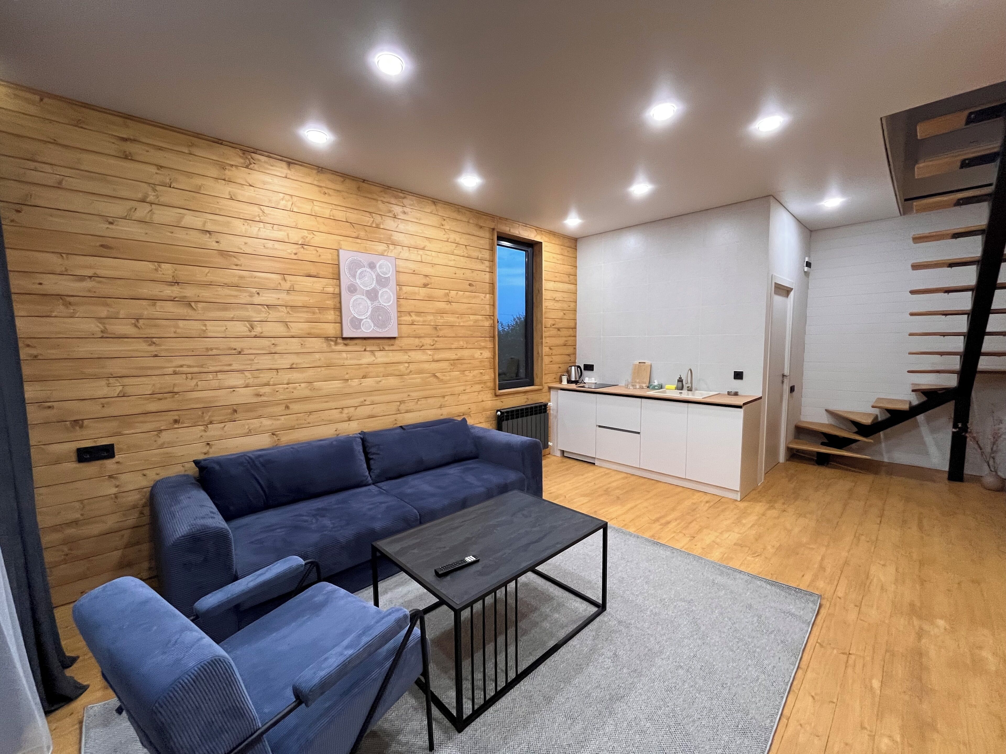 Un espacio habitable moderno con pared de madera, un sofá azul, una mesa de centro, zona de cocina y una escalera.