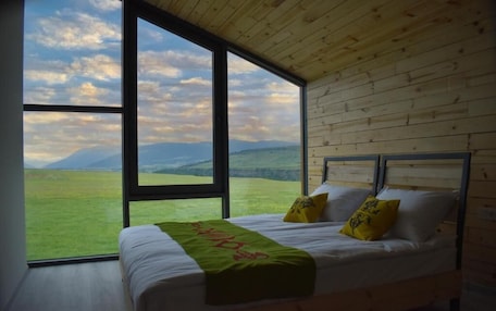 A bedroom with a large window, wooden walls, a bed with white bedding and two pillows, and a green throw blanket.