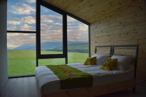 Un dormitorio con un gran ventanal, paredes de madera, una cama con ropa blanca y dos almohadas, y una manta verde.