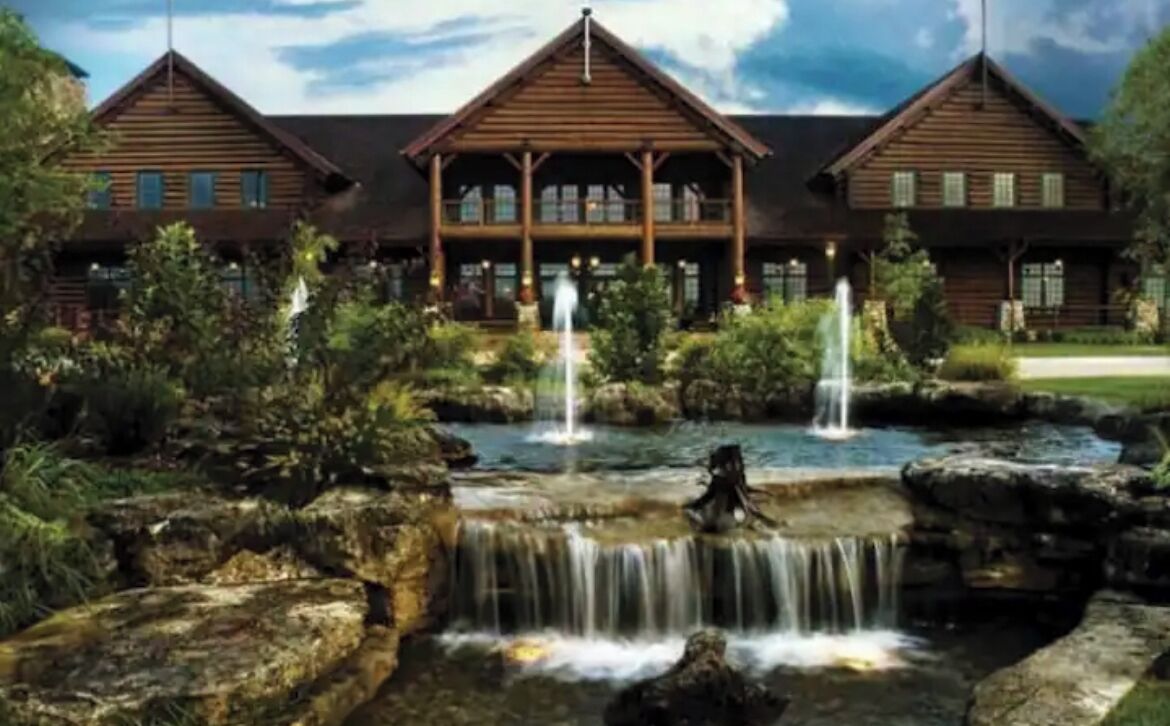 A large lodge with a central building and two smaller wings, surrounded by a natural landscape with a waterfall and rock formations.