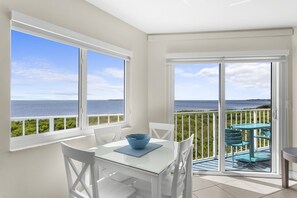 A room with a table and chairs, a view of the sea through large windows, and a balcony with a table and chairs.
