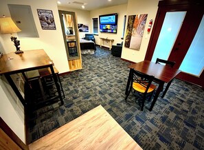 A hotel room with a large TV, a wooden table, a chair, a sofa, and a patterned carpet.