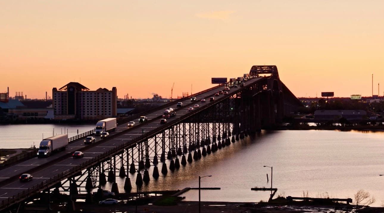 A bridge with multiple lanes of traffic, a body of water below, and industrial buildings in the background.