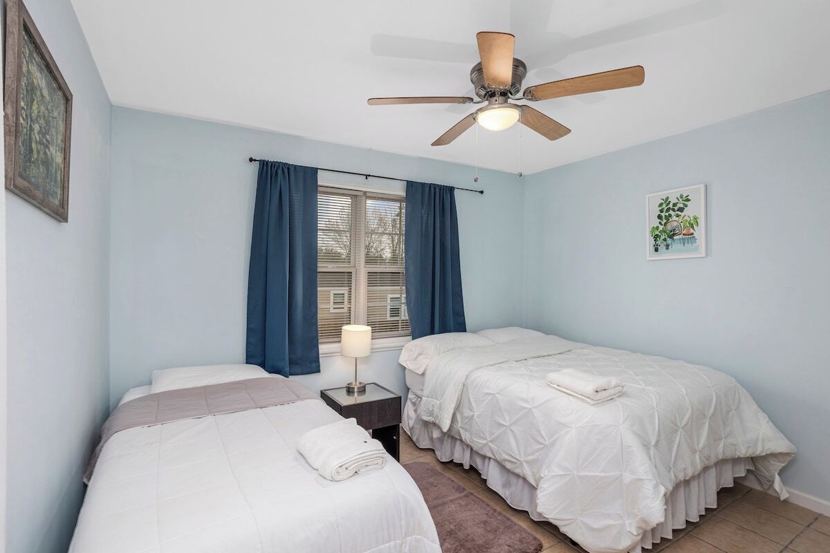 A bedroom with two beds, a ceiling fan, a window with curtains, and a bedside lamp.