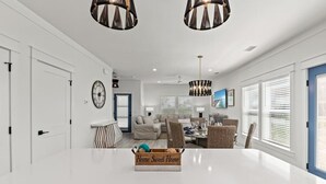 Una cocina moderna con encimeras blancas, un comedor con sillas de mimbre y una sala de estar con sofá y televisión.