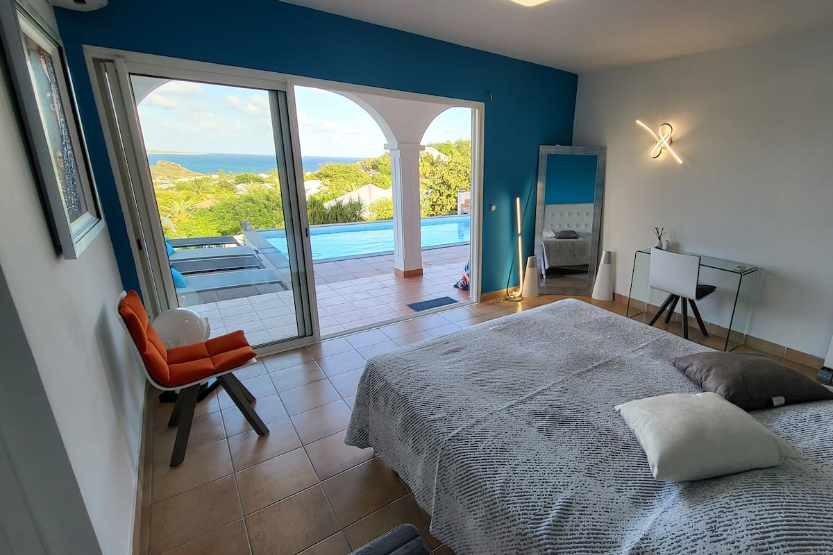 Ett sovrum med en stor säng, en stol, ett skrivbord och utsikt över en swimmingpool och grönområden.