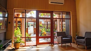 A modern room with a large sliding glass door leading to an outdoor area with plants and a thatched roof structure. There is a built-in air conditioning unit above the door, and the room has a wooden floor and two grey armchairs.