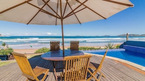 Une terrasse en bois avec une table et des chaises, un parasol et une vue sur l’océan et la plage.